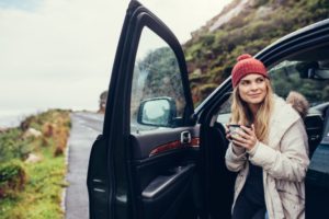 Woman standing outside of her car in winter clothing holding a coffee mug